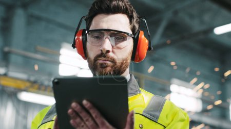 Photo for Bearded worker in protective headphones and googles wearing reflective yellow vest is looking at camera. Engineer standing and holding tablet in background of welding sparks flying. - Royalty Free Image