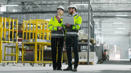Photo for Two chief engineers are discussing project and looking at laptop. Man and woman in yellow vests communicate at construction site. Employees gesturing on background of racks. - Royalty Free Image