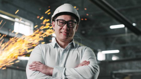 Photo for Asian male wearing white shirt, safety glasses and hard hat looks at camera. Engineer surrounded by equipment stands in background of work process with crossed armes and smiles. - Royalty Free Image