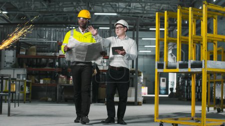 Photo for Two workers focused on their work walk around shop. Engineers in protective clothes. African-American employee holds blueprint in hands. Asian man is holding tablet and explaine something to colleague - Royalty Free Image