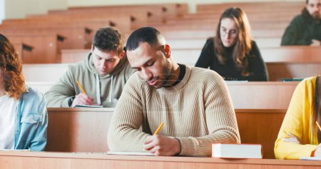 Téléchargez les photos : Jeune étudiant afro-américain beau garçon écoutant lection à l'université, écrivant et utilisant le manuel. - en image libre de droit