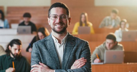 Téléchargez les photos : Portrait d'enseignant masculin heureux caucasien dans des lunettes se tournant vers la caméra et souriant joyeusement à l'université. Portrait de l'homme professeur en classe. Chargé de cours avec dossier de papiers. Les étudiants en arrière-plan. - en image libre de droit