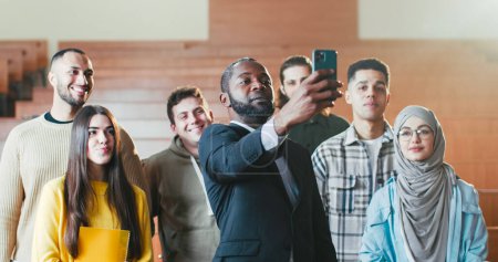 Téléchargez les photos : Afro-Américain professeur heureux de prendre des photos selfie avec smartphone parmi les étudiants métis en salle de classe de l'université. Concept éducateur et élèves. Faire des photos avec le téléphone. - en image libre de droit