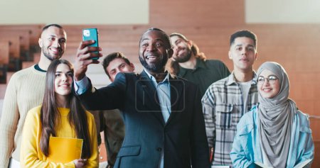 Téléchargez les photos : Portrait de l'homme afro-américain professeur joyeux parmi les étudiants multiethniques dans l'auditorium de l'Université riant et s'amusant devant la caméra. Concept éducateur et élèves. Amusant au lycée. - en image libre de droit