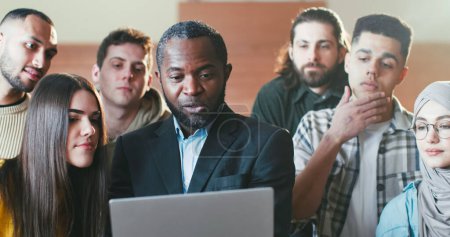 Téléchargez les photos : Gros plan d'un professeur afro-américain avec ordinateur portable parlant avec des étudiants multiethniques et montrant quelque chose à l'écran. Concept universitaire. Homme enseignant expliquant la matière à l'ordinateur. - en image libre de droit