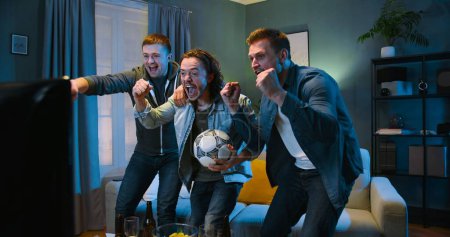 Tensed worried Caucasian male football fans friends staying in front of TV screen in dark living room on vouch and watching match Euro2024.Cheerful men celebrating win.