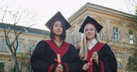 Téléchargez les photos : Portrait de jeunes filles multiethniques meilleures amies, heureuses diplômées d'étudiants universitaires en casquettes académiques et robes avec des diplômes dans les mains debout et souriantes. - en image libre de droit