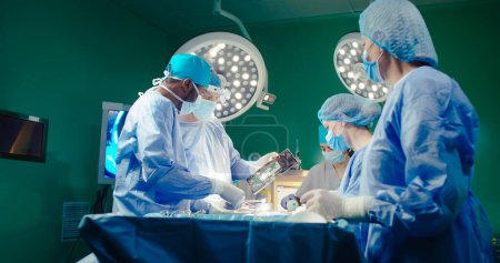 Vielfältiges Team professioneller Chirurgen, die invasive Operationen am Patienten im Operationssaal des Krankenhauses durchführen. Afroamerikanische Assistentin verteilt Instrumente an Chirurgen. Ethnische Vielfalt.