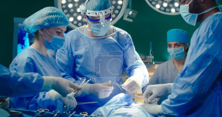 Professioneller Chirurg näht Patientin im Operationssaal. Eine Gruppe von medizinischem Personal in speziellen Uniformen, Handschuhen und Masken führt die Operation mit Hilfe moderner Ausrüstung aus. Soforthilfe.