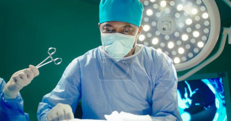 Photo for African American doctor close up. Doctor in special cap and protective mask. Doctor on background of surgery room. Doctor give scissors to assistant. Medical workers perform operation. - Royalty Free Image