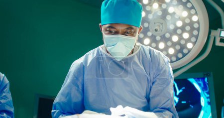 Photo for African American doctor close up. Doctor in special headdress and mask. Doctor on background of operating lamp. Doctor in gloves holds scissors and performs operation. Medical worker at workplace. - Royalty Free Image