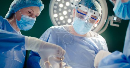 Photo for Operation, doctors dressed up in special clothes. Male doctor in binoculars. African American in special mask and gloves asssists operation. Medical workers at background of operating lamp. - Royalty Free Image