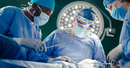 Trois travailleurs médicaux à l'arrière-plan de la lampe chirurgicale. Le médecin en chef en jumelles prend des ciseaux. Assistant afro-américain en salopette tenant des ciseaux. Une stagiaire aide à l'intervention.