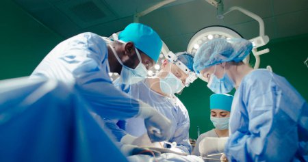 Tirez par en bas. Médecins en salopette en salle d'opération. Un Afro-Américain donne des ciseaux au docteur. Les internes féminines assistent pendant le processus chirurgical. Médecin en jumelles coud la plaie.