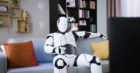Humanoid robot sitting on comfy couch and holding remote control in hand in living room. Futuristic white cyborg switching channels on TV set at home.