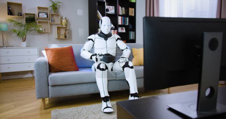 Robotic droid relaxing on cozy couch and watching show entertaining programs alone at home. Bionic white cyborg surprising with new information in video on television.