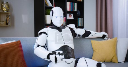 Retrato de humano como robot relajándose en un sofá gris en la sala de estar. Cyborg moderno futurista usando control remoto para cambiar canales de TV en apartamento con interior moderno.