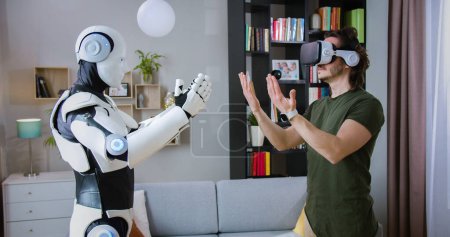 Seitenansicht einer menschenähnlichen Robotermaschine, die alle Bewegungen des unrasierten Menschen in VR-Brillen im Wohnzimmer wiederholt. Konzept von Menschen, Innovation und künstlicher Intelligenz.