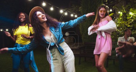 Joyful beautiful mixed-race girls dancing in garden at party. Caucasian and African American females having fun outdoor. Happy guy moving to music. Gathering together on summer evening Friends concept