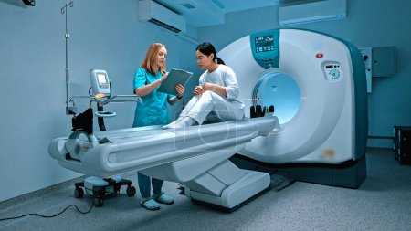Doctor holding folder and making notes of patient symptoms. Patient lying at CT scanner. Modern equipped cabinet in private clinic. Doctor talking to patient before procedure. MRI examination.