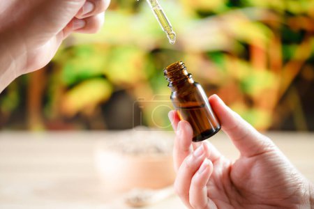 Hands holding a bottle of CBD oil and its dropper lid, with hemp leaf in the background. Legalized CBD product for medical purposes.