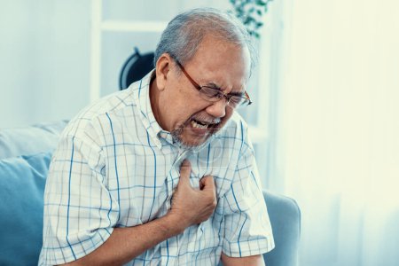Photo for An agonizing senior man suffering from chest pain or heart attack alone in his living room. Serious health problem and feeling unwell concept for seniors. - Royalty Free Image