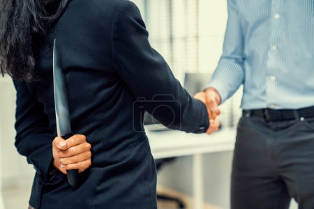 Foto de Back view of businesswoman shaking hands with another businessman while holding a knife behind his back. Concept of back backstabbing in business, backstabbing between colleagues. - Imagen libre de derechos