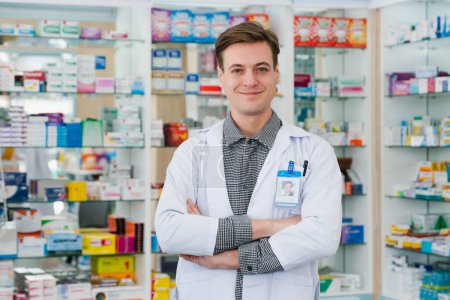 Portrait of a qualified, friendly male pharmacist wearing a white coat, crossing his arms, and looking at the camera, with a shelf of various medicine boxes in background at drugstore or pharmacy.