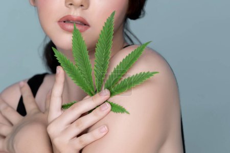 Foto de Closeup portrait of charming girl with fresh skin holding green leaf for beauty skin care made from cannabis leaf. Cosmetology and cannabis concept with isolated background. - Imagen libre de derechos