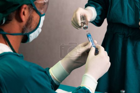 Photo for Surgeon fill syringe from medical vial for surgical procedure at sterile operation room with assistance nurse. Doctor and medical staff in full protective wear for surgery prepare anesthesia injection - Royalty Free Image