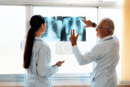 In a hospital sterile room, two professional radiographers hold and examine a radiograph for medical xray diagnosis. Novice doctor seeks advice on a patients condition from experienced older doctor.