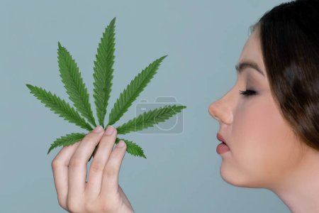 Foto de Closeup charming girl with fresh skin hold green leaf for beauty skin care made from cannabis leaf. Cosmetology and cannabis concept with isolated background. Woman holding cbd leaf side view. - Imagen libre de derechos