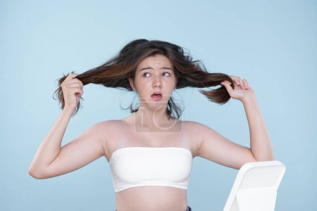 Foto de Beautiful girl feel sad, worry about her damaged hair with isolated background. Concept of dry lifeless hair. Young charming girl hold her tangled hair and look at it with worried expression. - Imagen libre de derechos