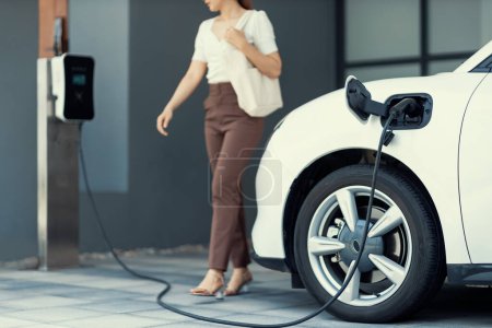 Foto de Focus image of electric vehicle recharging battery at home charging station with blurred woman walking in the background. Progressive concept of green energy technology applied in daily lifestyle. - Imagen libre de derechos