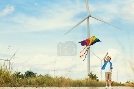 Photo for Progressive happy carefree boy concept. Young boy running along the road and flying kite at natural scenic on countryside, clear sky and sunny day with mountain and wind turbine background - Royalty Free Image