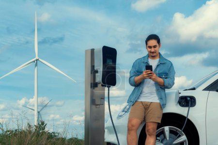 Téléchargez les photos : Homme progressiste avec sa voiture électrique, la voiture EV recharge l'énergie de la station de recharge sur champ vert avec éolienne comme concept d'énergie durable future. Véhicule électrique avec générateur d'énergie. - en image libre de droit