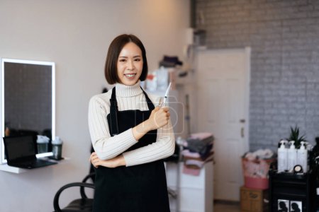 Téléchargez les photos : Portrait d'une jeune coiffeuse asiatique tenant des outils de coupe de cheveux qualifiés dans son salon pour une coupe de cheveux de femmes. Concept d'emploi photo pour propriétaire de petite entreprise et soins capillaires. - en image libre de droit
