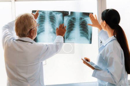 Téléchargez les photos : Dans une salle stérile de l'hôpital, deux radiologistes professionnels tiennent et examinent une radiographie aux fins de diagnostic par rayons X médicaux. Novice médecin demande conseil sur un état des patients auprès d'un médecin plus âgé expérimenté. - en image libre de droit