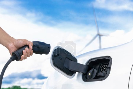 Progressive natural scenic with windmill generator where hand insert charging plug to electric vehicle from charging station with natural background. EV car powered by wind turbine electric generator.