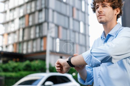 Téléchargez les photos : Businessman with smartwatch at modern charging station for electric vehicle with background of residential buildings as concept for progressive lifestyle of using eco-friendly as alternative energy. - en image libre de droit
