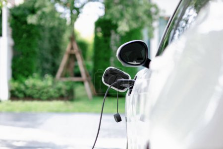 Foto de Closeup electric vehicle plugged-in with cable from charging point powered for progressive concept by alternative clean energy rechargeable EV car at home charging station. - Imagen libre de derechos