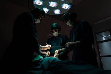 Foto de Equipo quirúrgico realizando cirugía al paciente en quirófano estéril. En una sala de cirugía iluminada por una lámpara, un equipo quirúrgico profesional y seguro proporciona atención médica a un paciente inconsciente. - Imagen libre de derechos