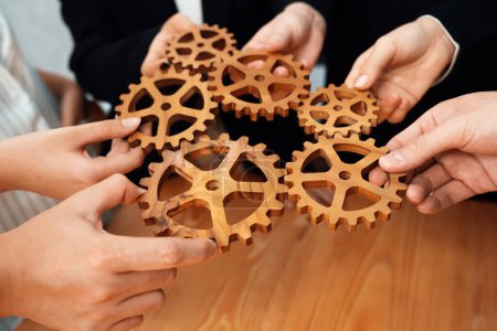 Téléchargez les photos : Closeup hand holding wooden gear by businesspeople wearing suit for harmony synergy in office workplace concept. Group of people hand making chain of gears into collective form for unity symbol. - en image libre de droit