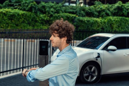 Téléchargez les photos : Businessman with smartwatch at modern charging station for electric vehicle with background of residential buildings as concept for progressive lifestyle of using eco-friendly as alternative energy. - en image libre de droit