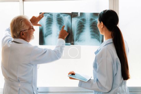 Photo for In a hospital sterile room, two professional radiographers hold and examine a radiograph for medical xray diagnosis. Novice doctor seeks advice on a patients condition from experienced older doctor. - Royalty Free Image