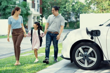 Foto de Progressive young happy family with electric vehicle and home backyard charging station. Green and clean energy from electric vehicles for healthy environment. Eco power from renewable source. - Imagen libre de derechos