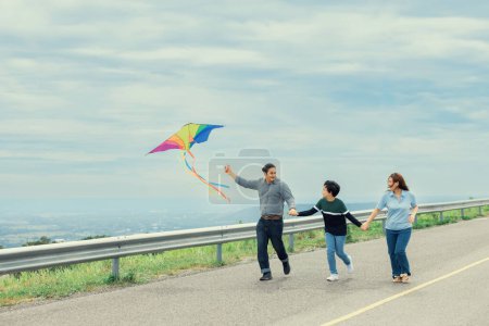 Photo for Progressive happy family vacation and carefree day concept. Young parents mother father and son run along and flying kite together road with enjoy natural scenic on scenery and clear sky background. - Royalty Free Image