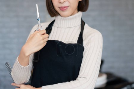 Téléchargez les photos : Portrait d'une jeune coiffeuse asiatique tenant des outils de coupe de cheveux qualifiés dans son salon pour une coupe de cheveux de femmes. Concept d'emploi photo pour propriétaire de petite entreprise et soins capillaires. - en image libre de droit