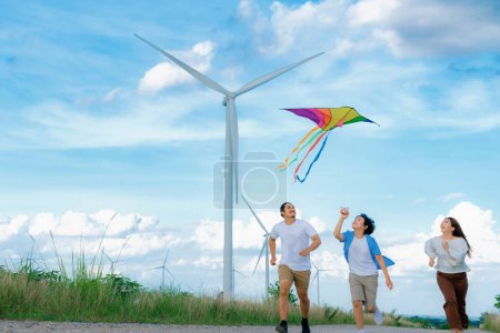 Photo for Progressive happy carefree family vacation concept. Young parents mother father and son run along and flying kite together on road with natural scenic on mountain and wind turbine background. - Royalty Free Image