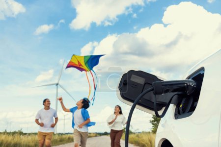 Téléchargez les photos : Concept of progressive happy family holding windmill toy and relax at wind farm with electric vehicle. Electric vehicle driven by clean renewable energy from wind turbine generator to charger station. - en image libre de droit
